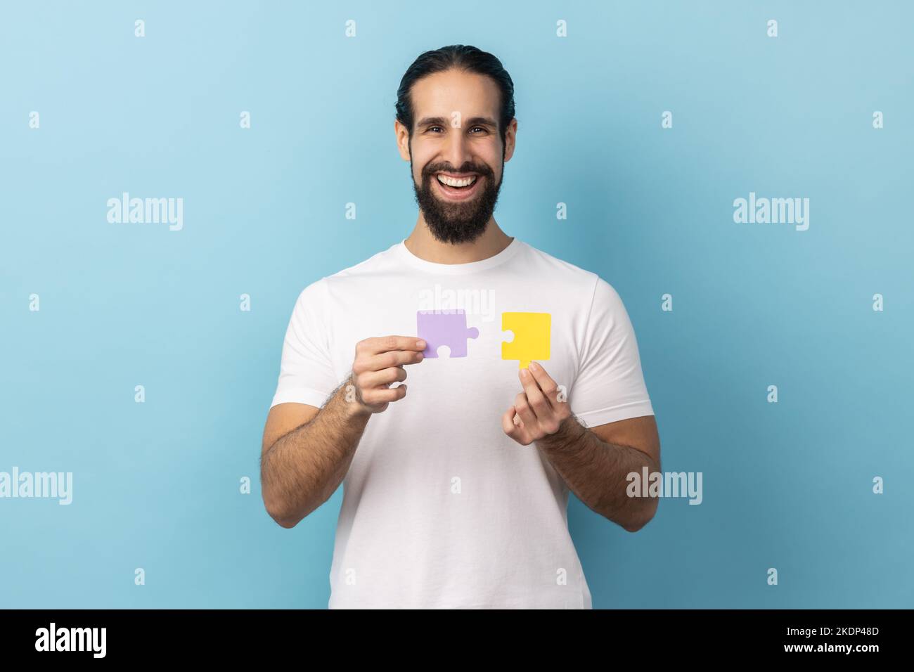Portrait of man with beard wearing white T-shirt holding yellow and purple puzzle pieces, solving tasks, looking at camera with toothy smile. Indoor studio shot isolated on blue background. Stock Photo