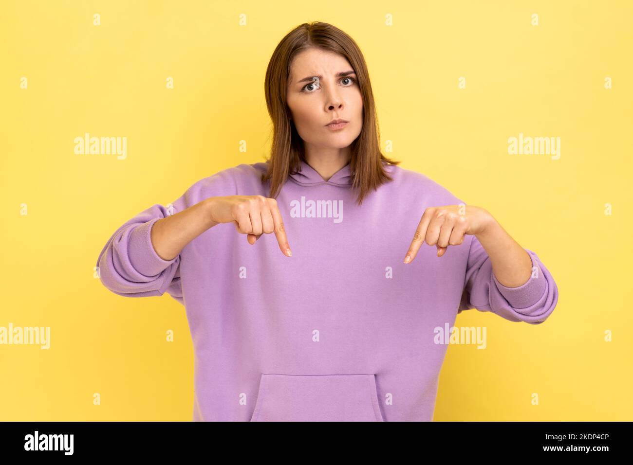 Portrait of serious strict confident bossy woman pointing fingers down, asking to act here and now immediately, wearing purple hoodie. Indoor studio shot isolated on yellow background. Stock Photo