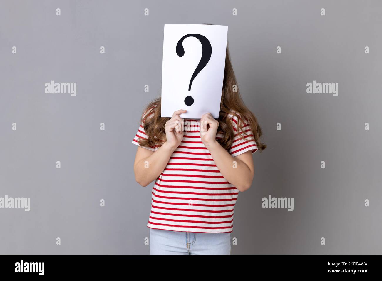Portrait of unknown little girl wearing striped T-shirt standing hiding face behind paper with question mark, having problem for solving. Indoor studio shot isolated on gray background. Stock Photo