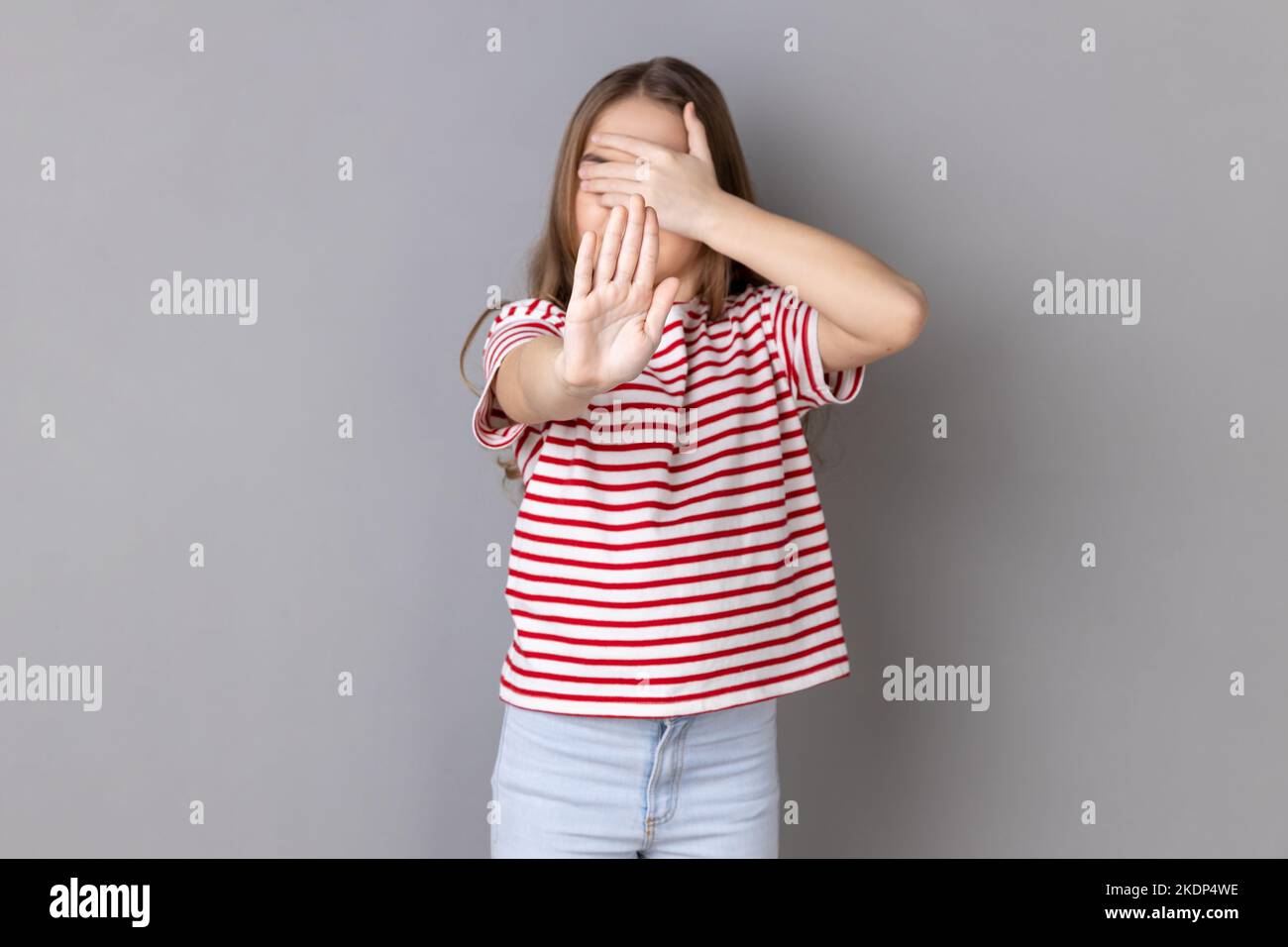Portrait of sacred little girl wearing striped T-shirt closing eyes with palm of hand, stretching out another hand forward, showing stop gesture. Indoor studio shot isolated on gray background. Stock Photo