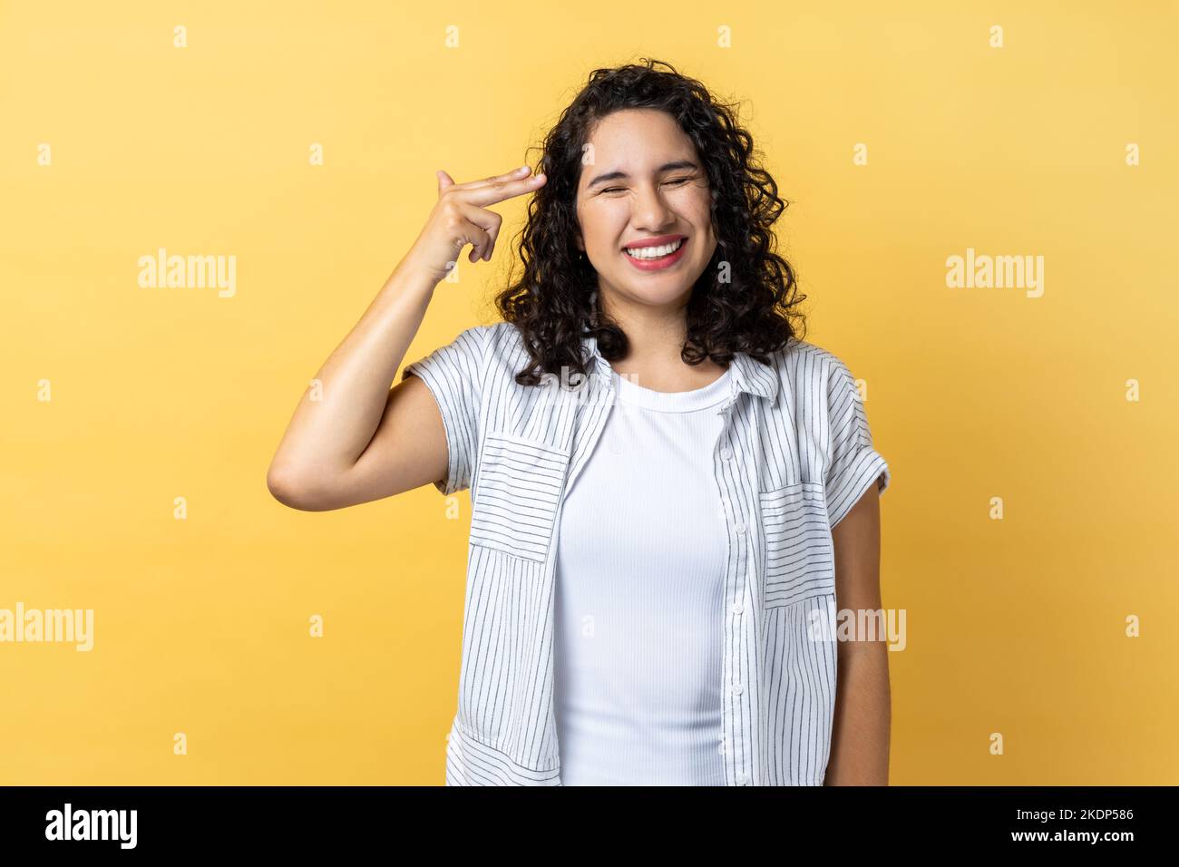 Portrait of woman with dark wavy hair pointing finger gun to head and looking desperate, making suicide gesture, shooting herself. Indoor studio shot isolated on yellow background. Stock Photo