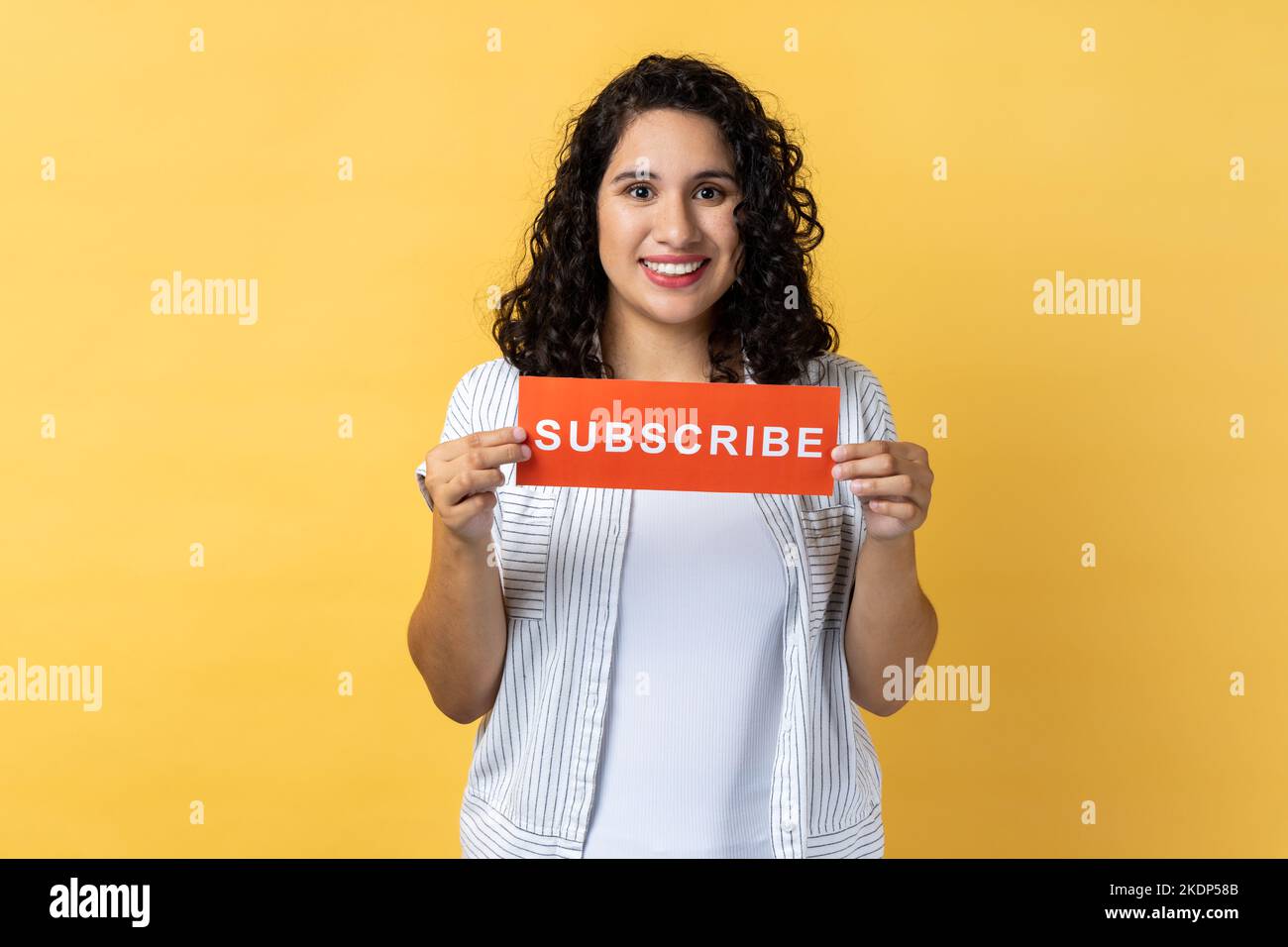 Portrait of happy smiling positive woman with dark wavy hair holding red card with subscribe inscription, asking to follow her blog. Indoor studio shot isolated on yellow background. Stock Photo