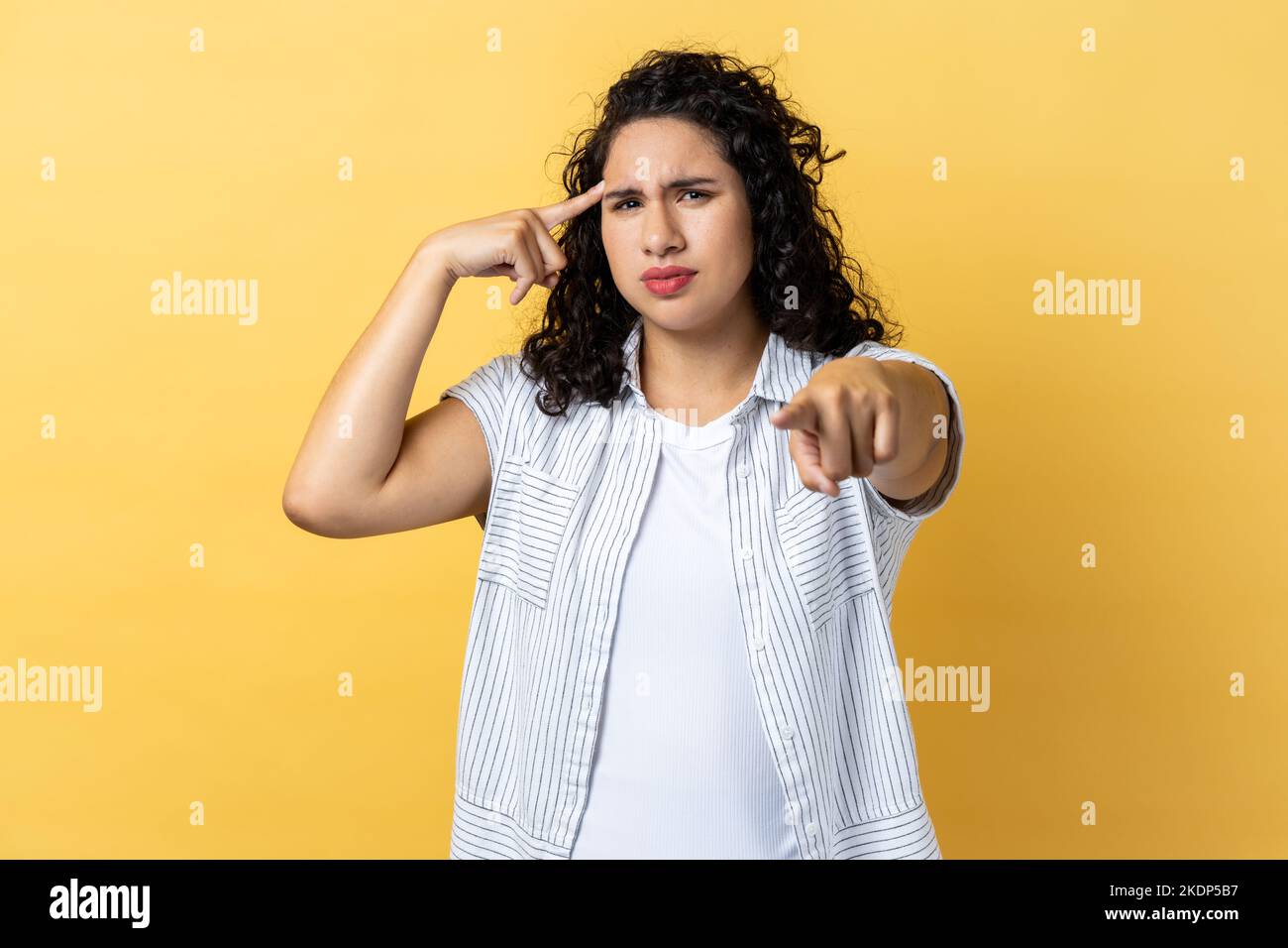 Portrait of winsome young adult woman with dark wavy hair holding finger near head and pointing finger at camera, stupid gesture. Indoor studio shot isolated on yellow background. Stock Photo