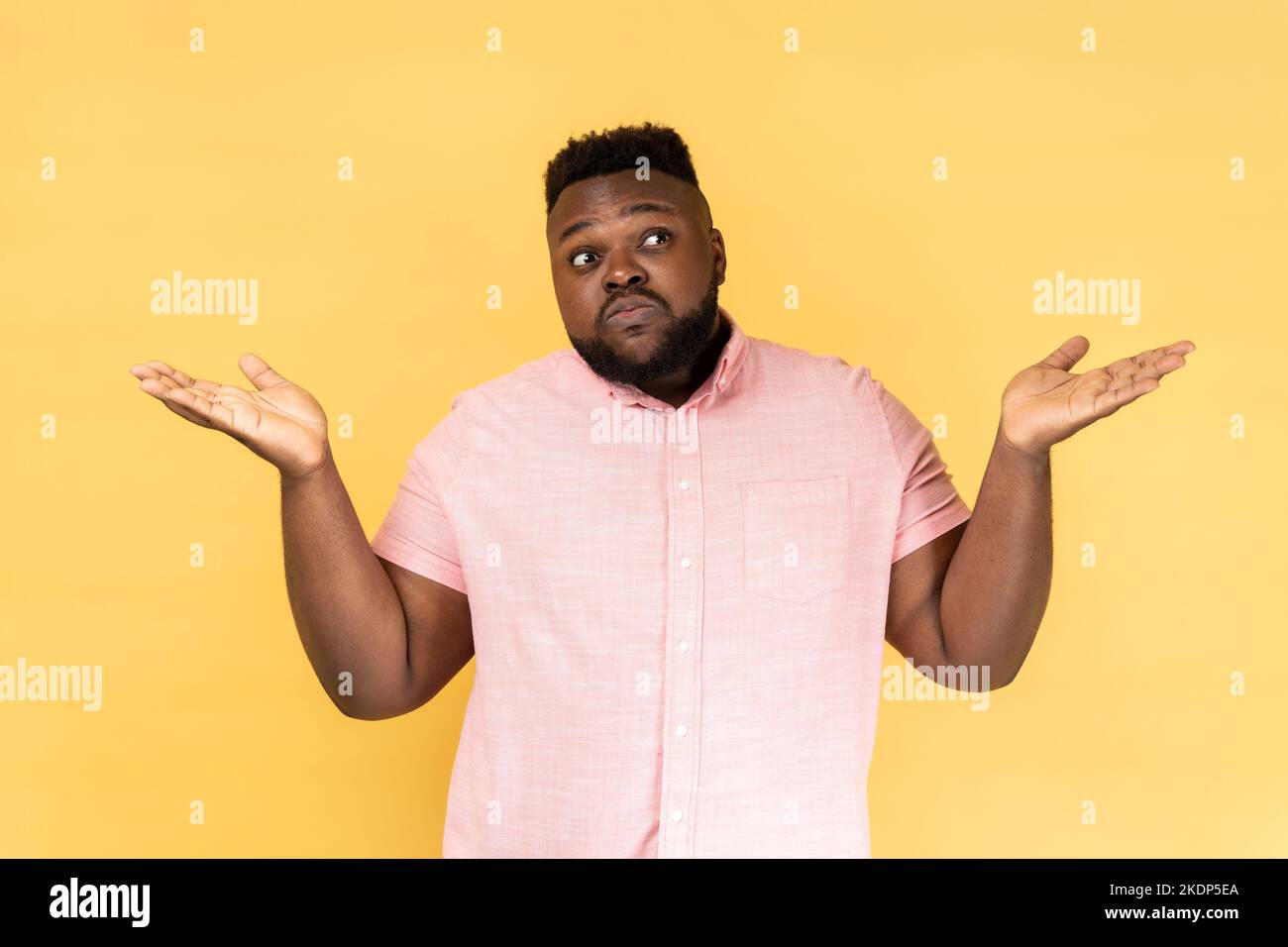I dont know. Portrait of confused man wearing pink shirt shrugging shoulders as doesnt know answer, cant make decision, being uncertain, not sure. Indoor studio shot isolated on yellow background. Stock Photo