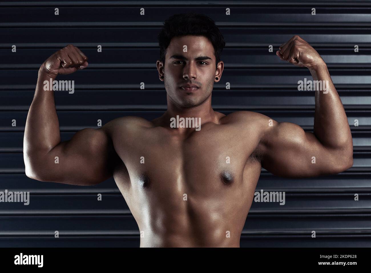 Check these guns. Cropped portrait of an athletic young man flexing his biceps against a dark background. Stock Photo