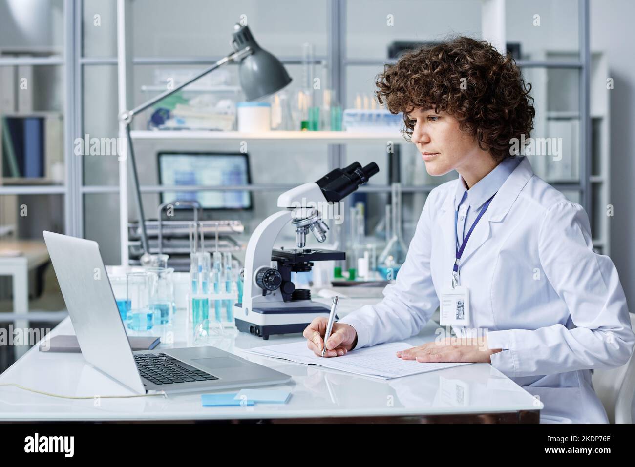 Young female scientist in labcoat making notes while sitting by workplace in front of laptop in laboratory and watching online video Stock Photo