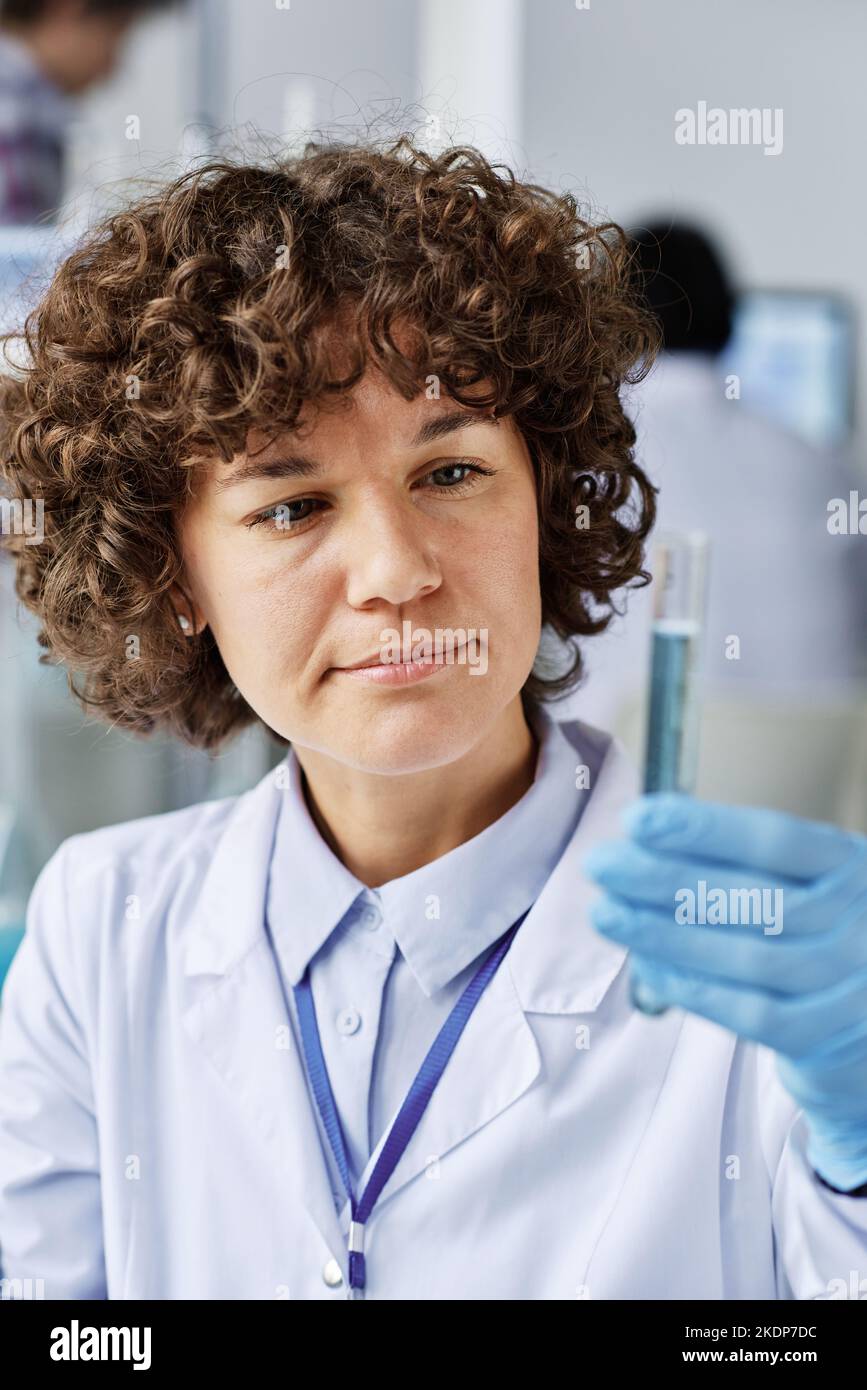 Young brunette woman in protective gloves and labcoat holding flask with liquid substance after mixing two fluids in laboratory Stock Photo