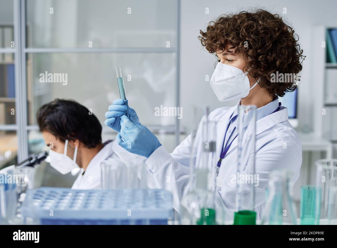 Young female scientist in respirator, labcoat and protective gloves looking at flask with blue liquid against colleague Stock Photo