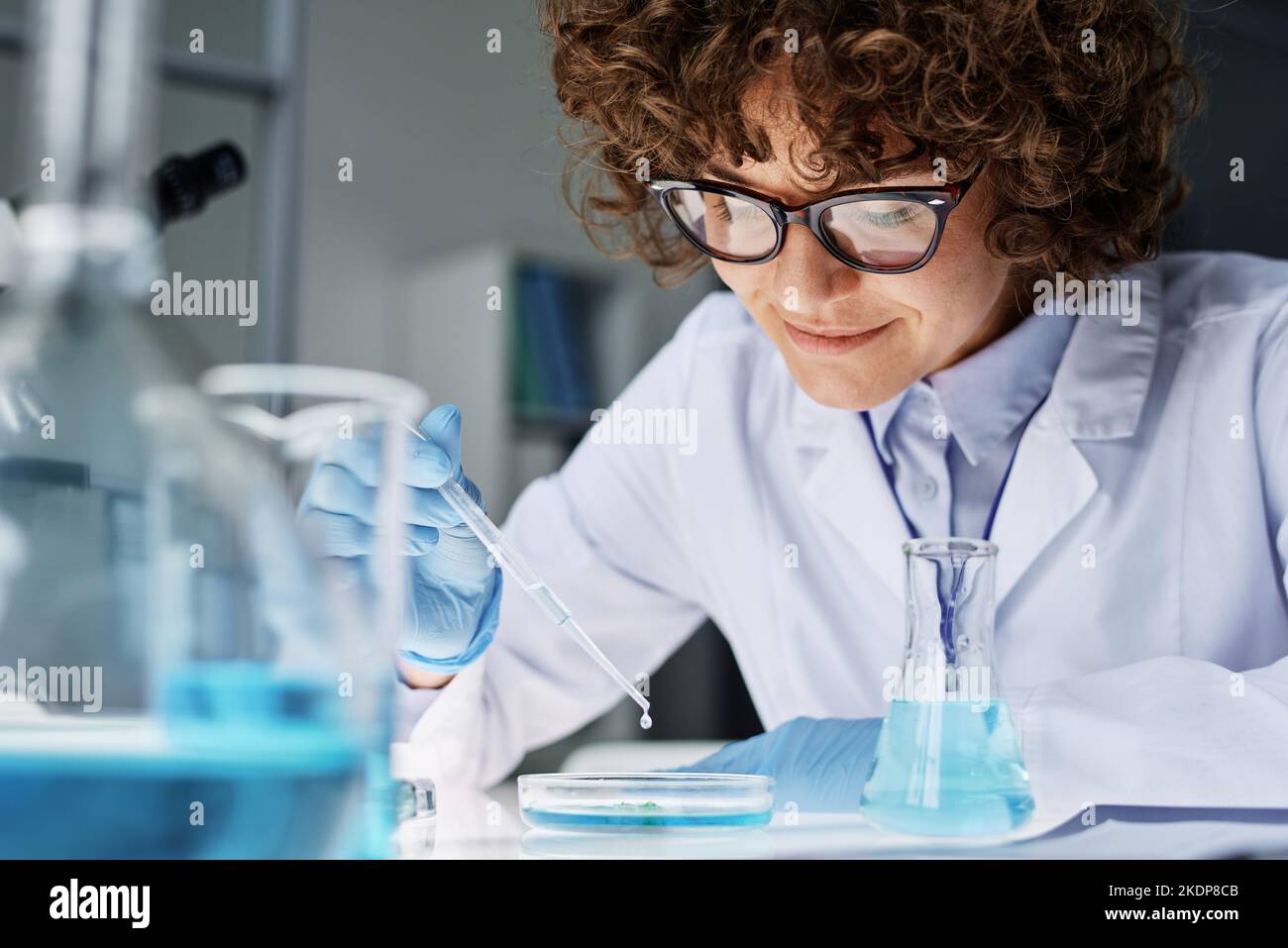 Young female scientist or chemist in labcoat, gloves and eyeglasses dropping liquid substance into petri dish by workplace Stock Photo