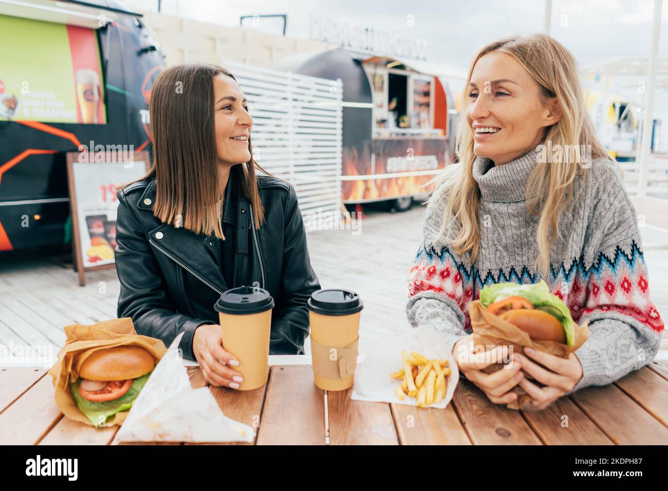 Two young modern european girlfriends are chatting and eating fast food at the street market. Stock Photo