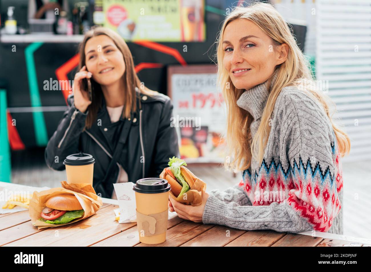 Two cute women eating burgers and chatting while sitting in the market near the food truck. Stock Photo