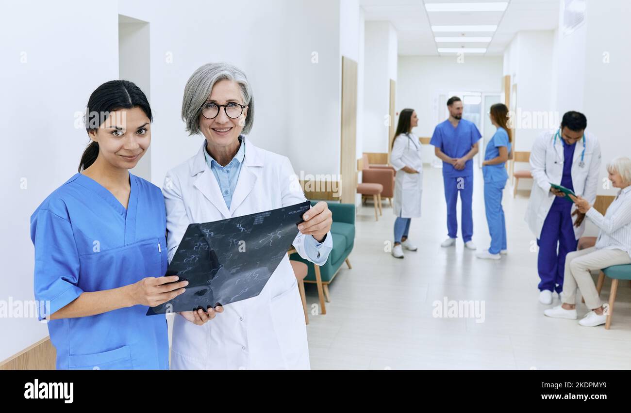 Female intern with her medical mentor in hospital corridor during medical practice against backdrop of doctors and medical staff. Internship and medic Stock Photo