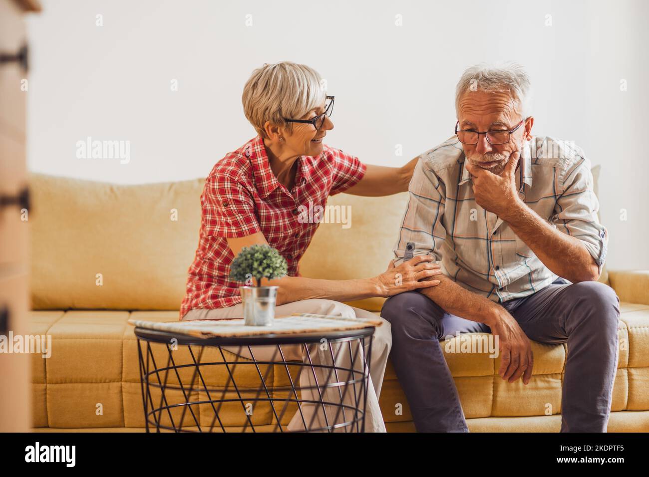 Senior couple sitting in living room. Man is sad and worried and woman is consoling him. Stock Photo