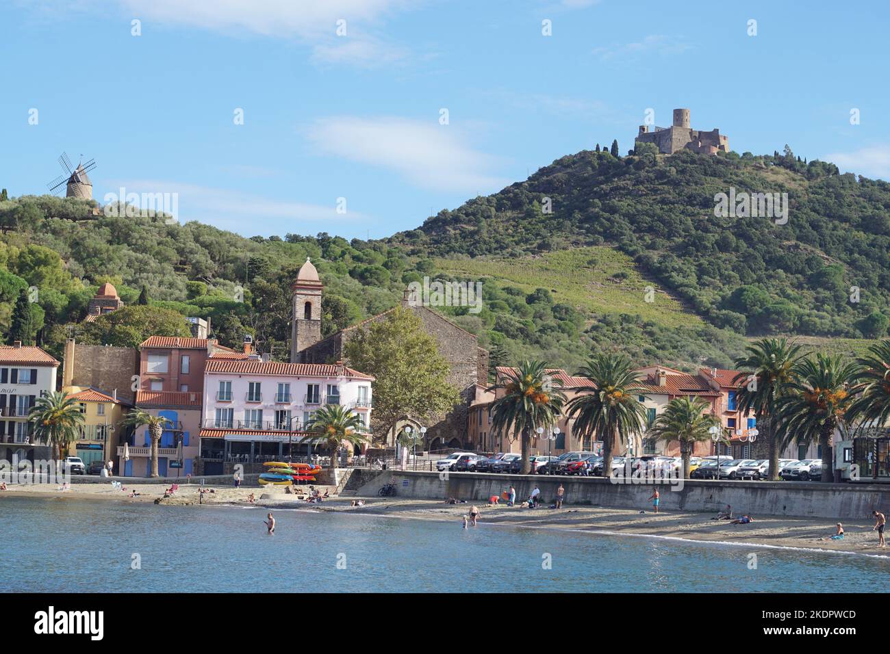 Collioure, France - October 2022; View across bay featuring people in water and on beach against backdrop of colourful buildings, and hill Stock Photo