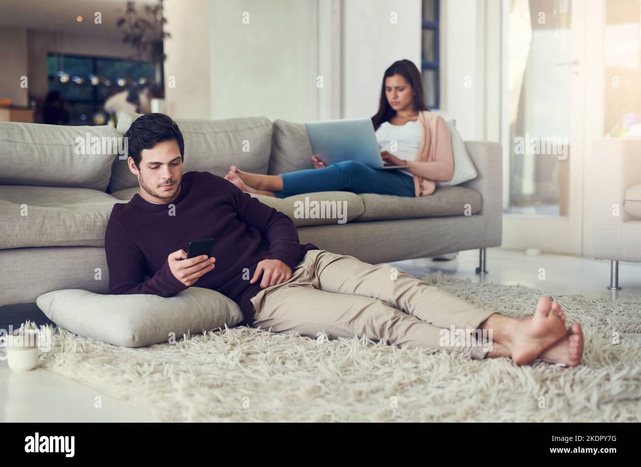 I am so glad that we can relax the whole day. a young couple relaxing in the living room while being on their mobile devices at home. Stock Photo