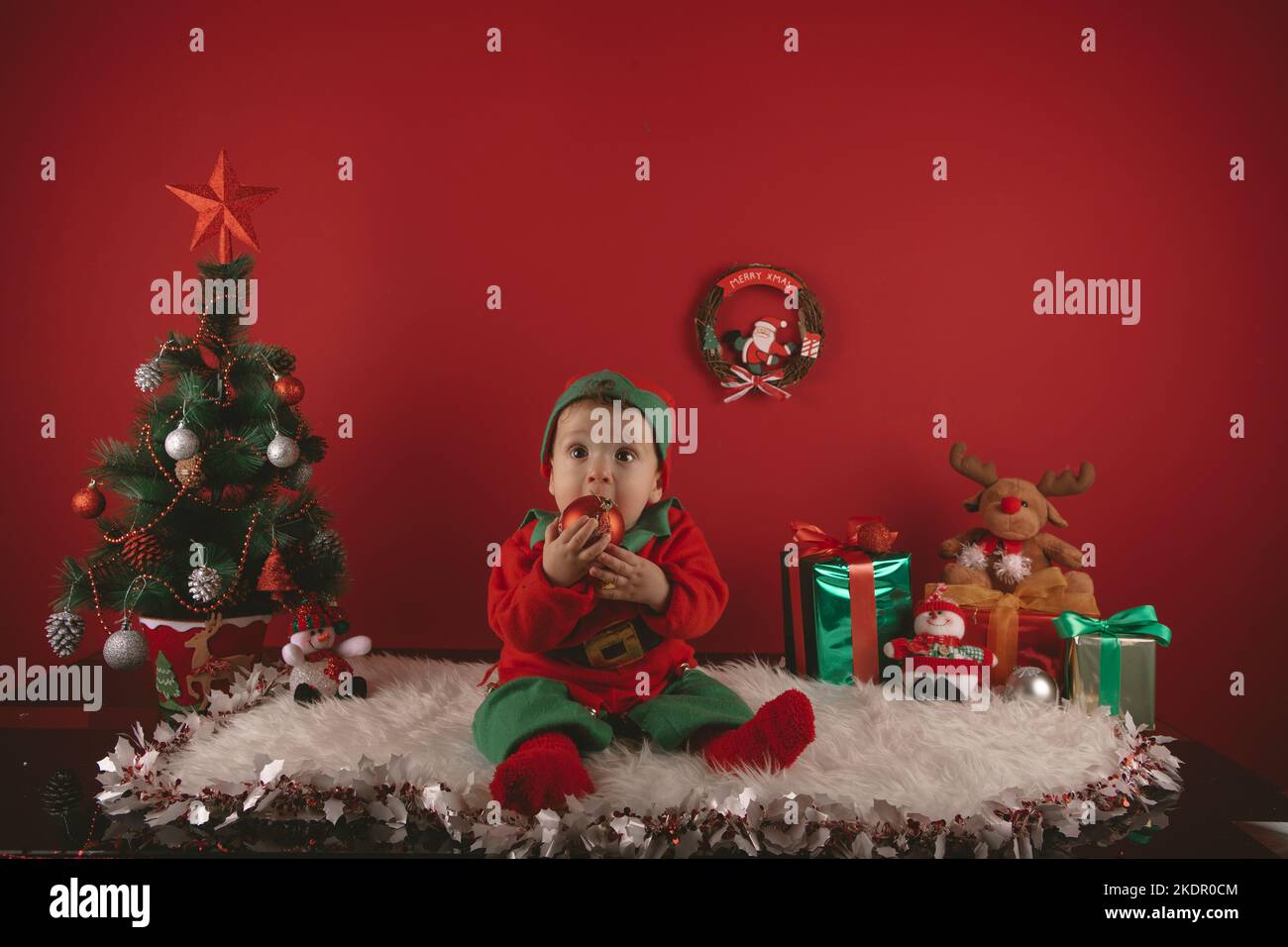 photo of a nice boy dressed as an elf next to a Christmas decoration with a tree and gifts Stock Photo