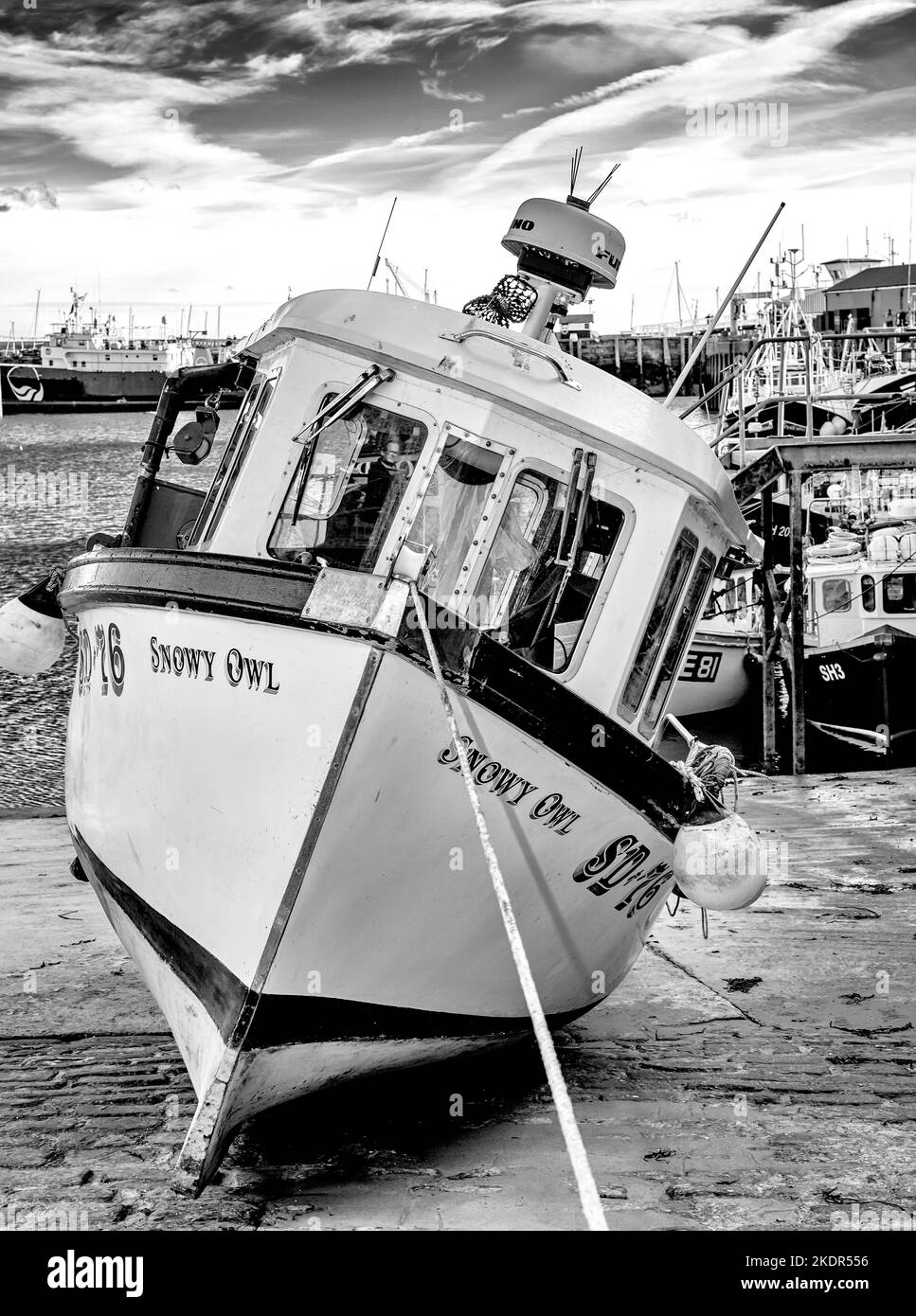 A boat lies to one side aground on a harbour slope. Fishing trawlers are beyond and a sky with cloud is above. Stock Photo