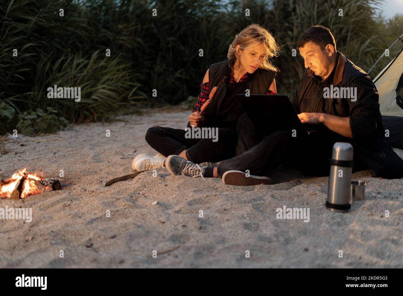 Freelancer working in a tent on a camping trip in the evening. Couple in love relaxing watching video at sunset on the beach in summer. Recreation Stock Photo