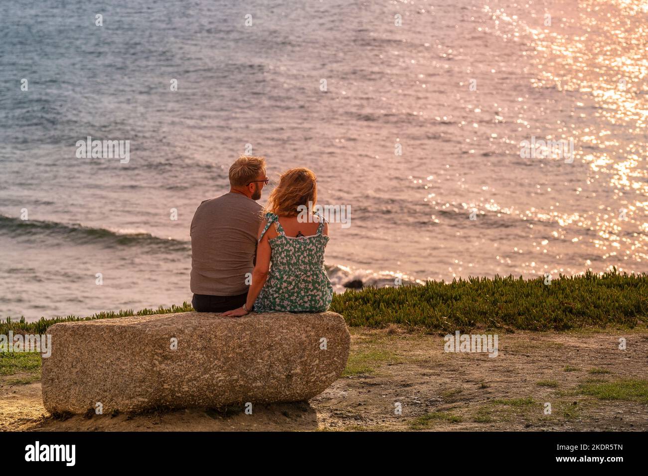 A couple sitting closely together on a rock on the coast overlooking Fistral Bay in Newquay in Cornwall in the UK in Europe. Stock Photo