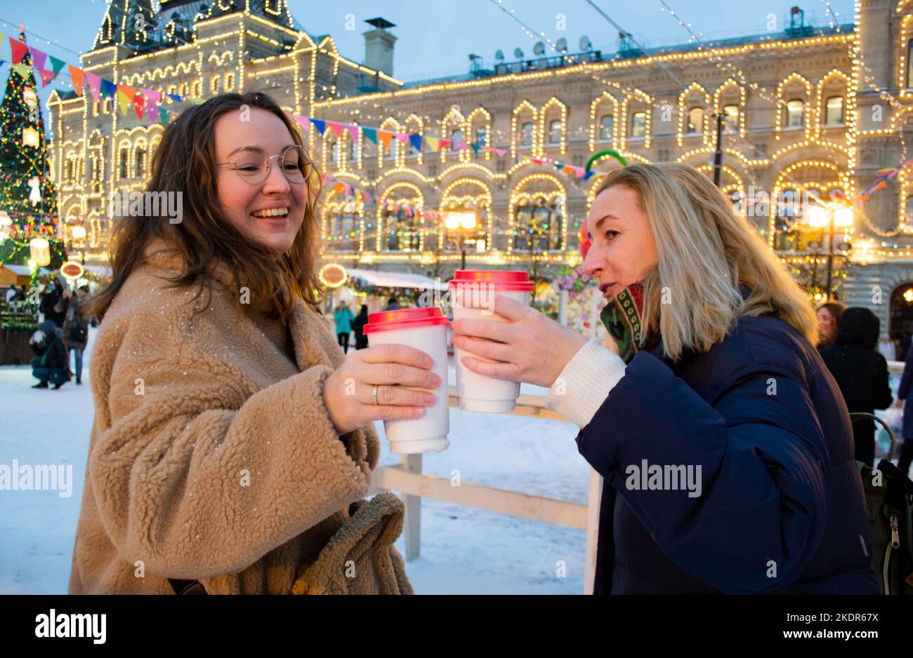 Two cheerful young women with cardboard cups of mulled wine cheers and laugh. Christmas fair on Red Square, Moscow. Stock Photo