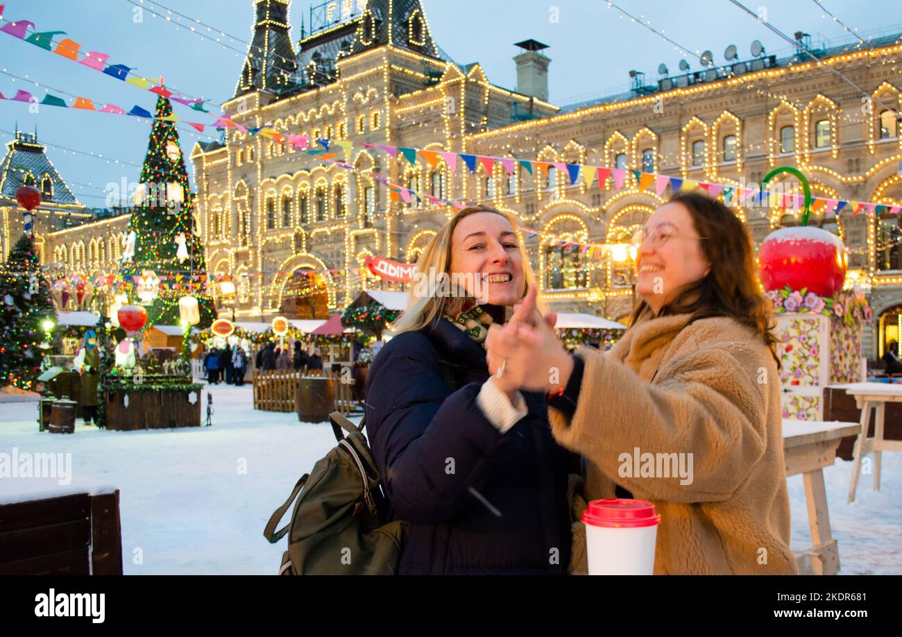 Two cheerful young women with cardboard cups with hot drinks dance and laugh. Christmas Fair in Moscow on Red Square. Stock Photo