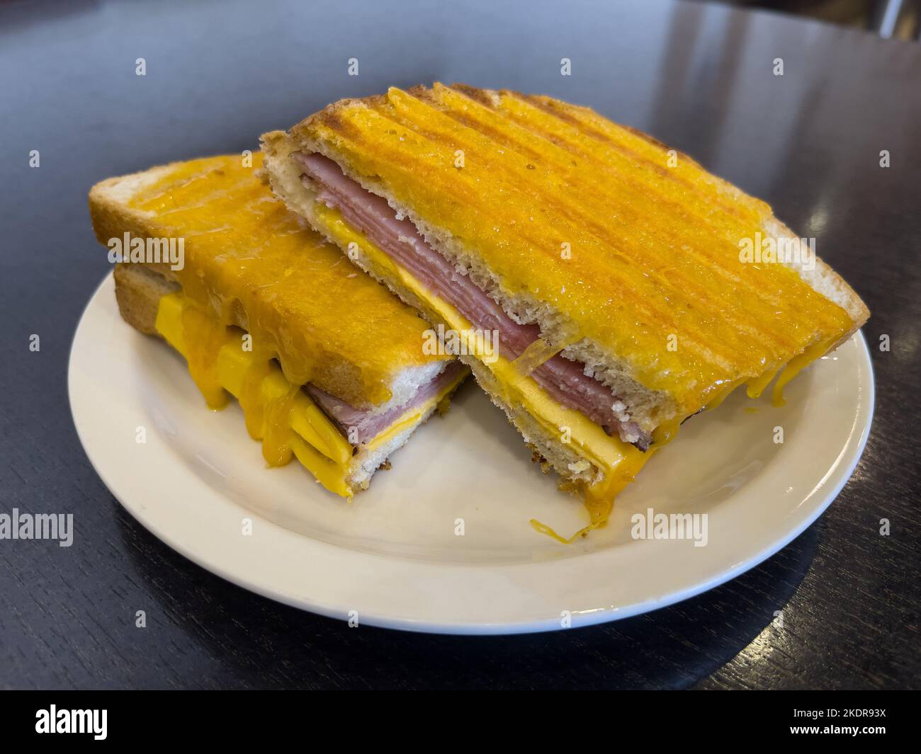 grilled ham and cheese sandwich served in a white plate and placed on a dark brown wooden table. Stock Photo