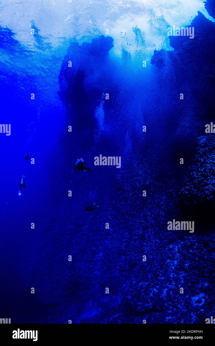 Group of divers in deep water next to steep wall and crashing waves above Stock Photo