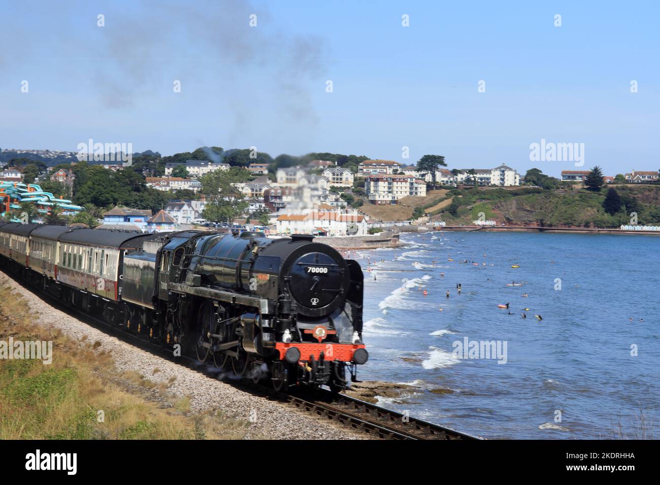 Class 7 Brittania 70000 steam loco as it passes Goodrington Sands, Paignton, with people in the sea enjoying a Summer day. Torbay, South Devon, Southw Stock Photo