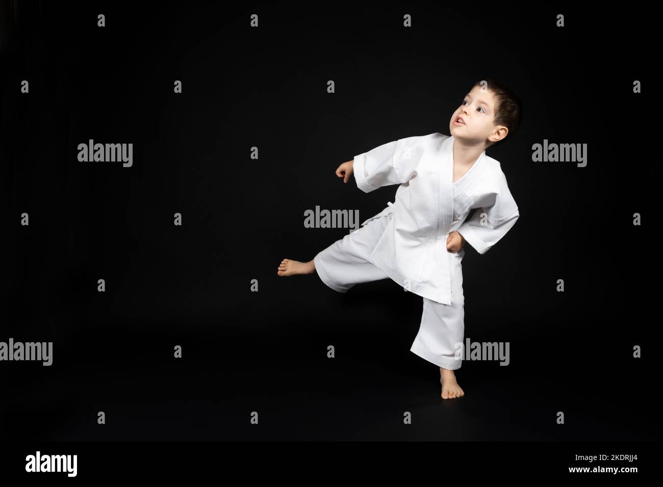 A little boy in a kimono practices karate on a black background, kicking forward. Stock Photo