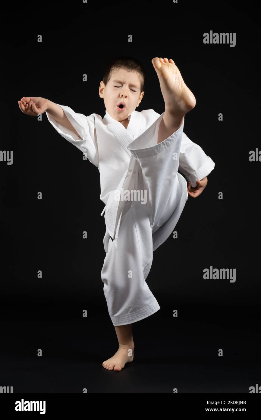 A little boy in a kimono practices karate on a black background, kicking forward. Stock Photo
