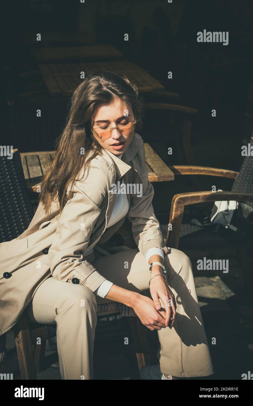 beautiful brunette girl in a sunglasses dressed in white blouse and beige coat posing in a chair. Stylish trendy fashion outlook Stock Photo