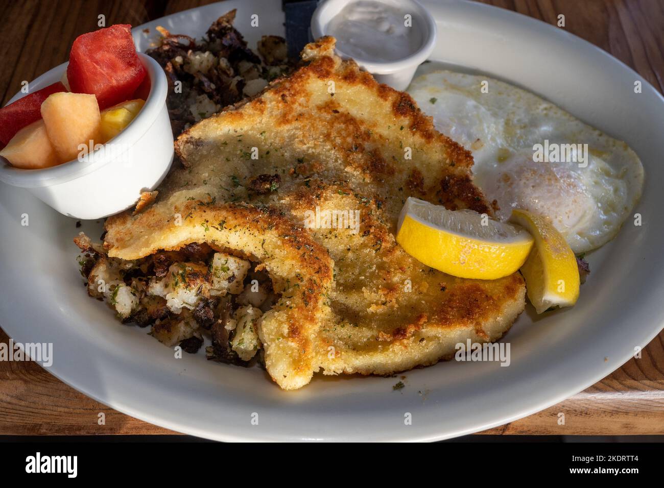 Seafood breakfast consisting of calamari steak, fried eggs, fruit and potato on white plate in a restaurant setting viewed from dinner angle Stock Photo