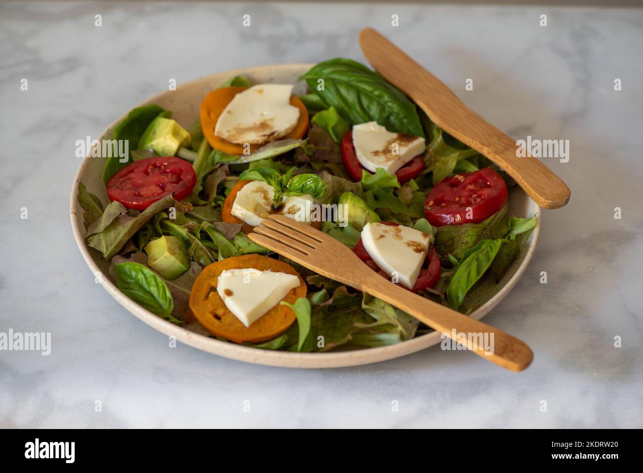 Organic mixed greens, tomatoes, avocado and mozzarella salad served with olive oil and balsamic vinegar in bamboo plate and silverware, on marble tabl Stock Photo