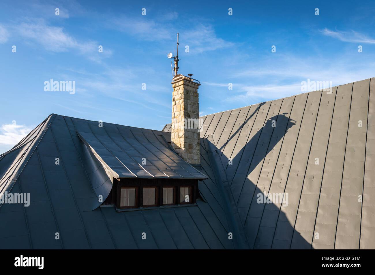 Hipped roof with roof window (dormer) and covered with grey metal seam sheet. Stone chimney with cap and little transmitter. Blue, cloudy sky in the b Stock Photo