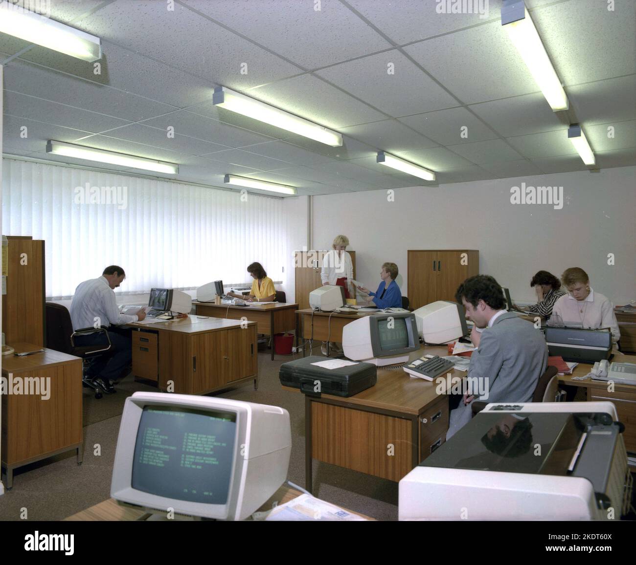 1980s, historical, view across an open office of the era, male and female employees working at desks, using small desk-top computer terminals, most likely made by IBM, England, UK. Being CRT monitors they displayed text in green, as seen in the picture and so became known as green screen terminals. Stock Photo