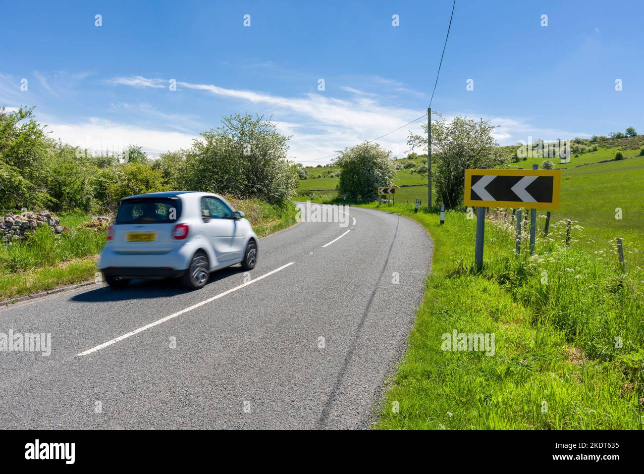 Traffic passing a black and white chevron road sign warning of an approaching left hand bend on Cliff Road at the top of Cheddar gorge in the Mendip Hills, Somerset, England. Stock Photo