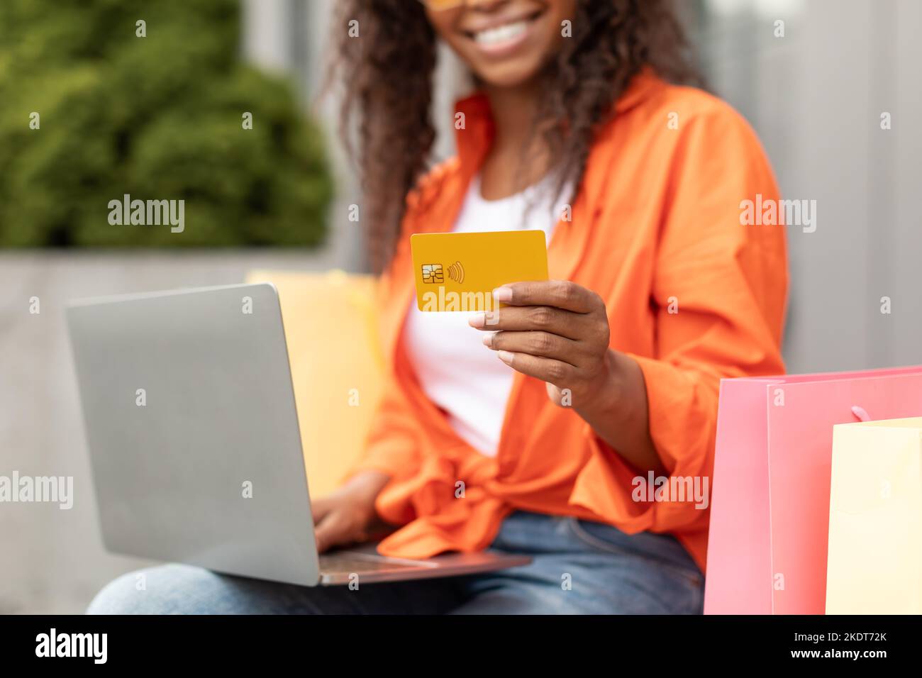 Smiling millennial black lady in casual with many bags typing on laptop, holding credit card in city, cropped Stock Photo