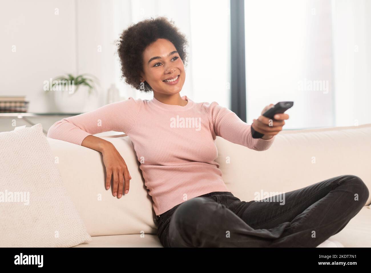 Smiling relaxed millennial african american woman with remote control watching tv sit on sofa, enjoy free time Stock Photo