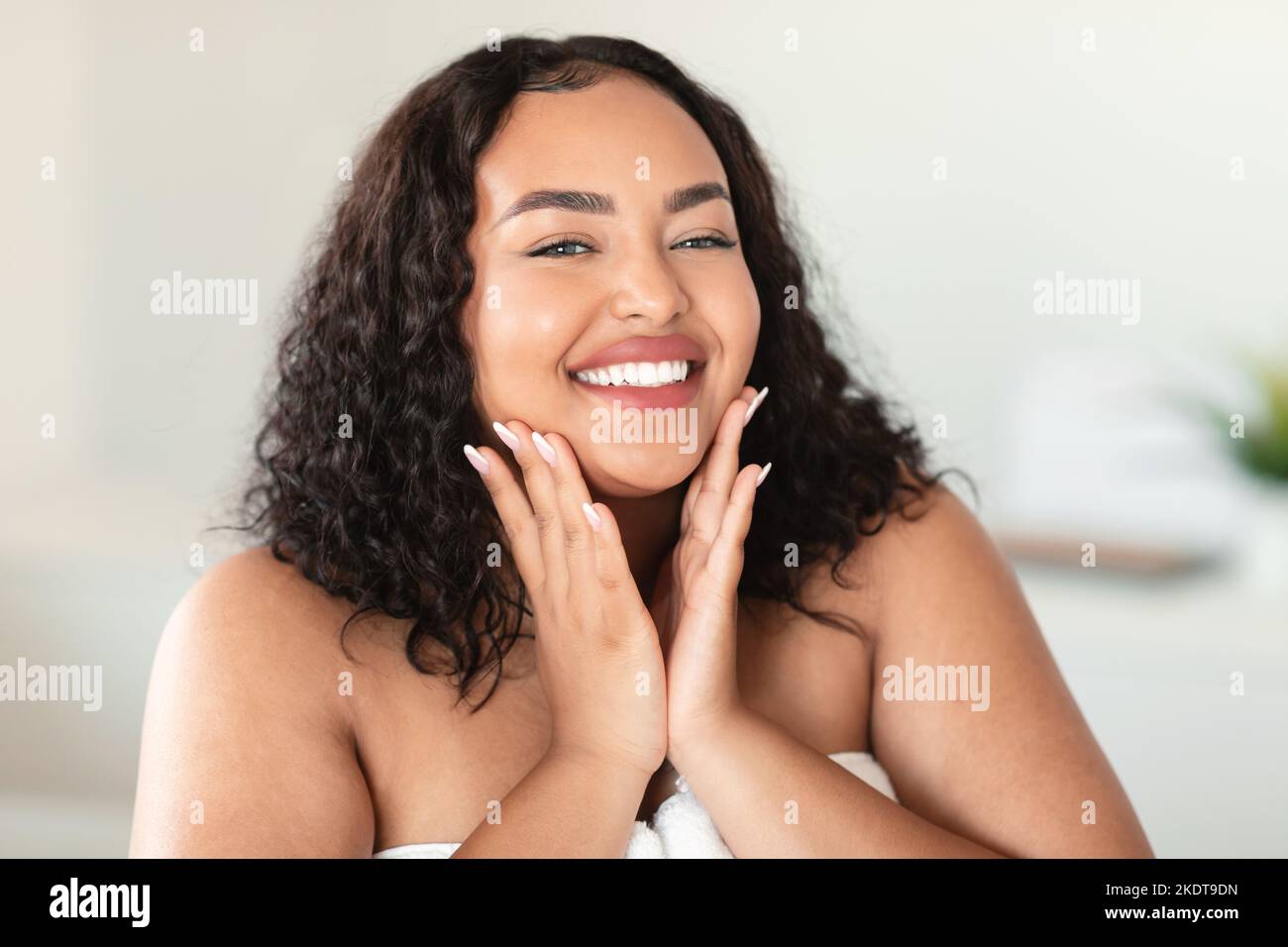 Portrait of beautiful black chubby woman touching her cheeks and smiling at camera, enjoying skincare treatments at home Stock Photo