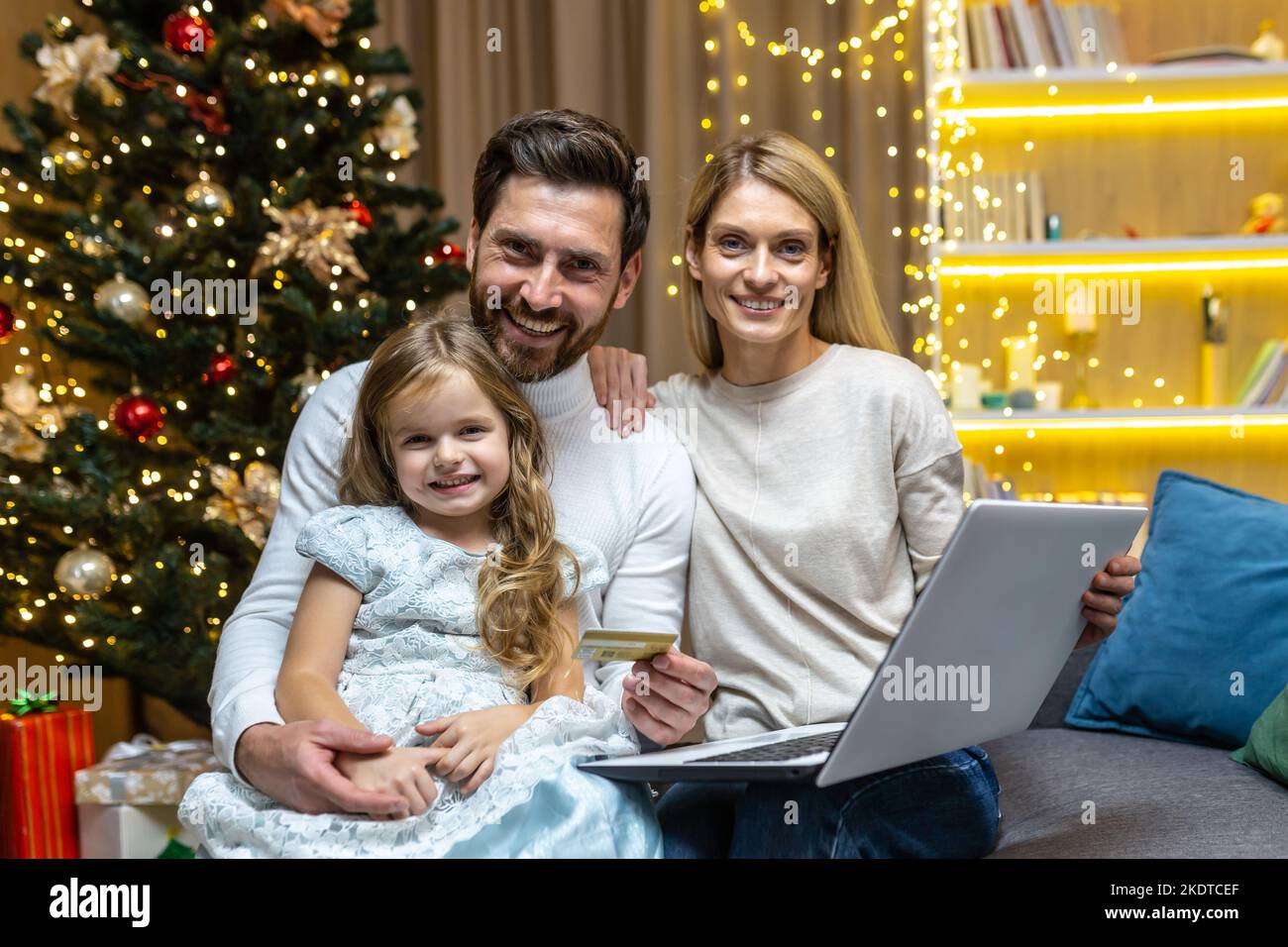 Portrait of a young family sitting on the sofa at home near the Christmas tree. Holding laptop and credit card, choosing Christmas gifts online. They look at the camera, smile. Stock Photo