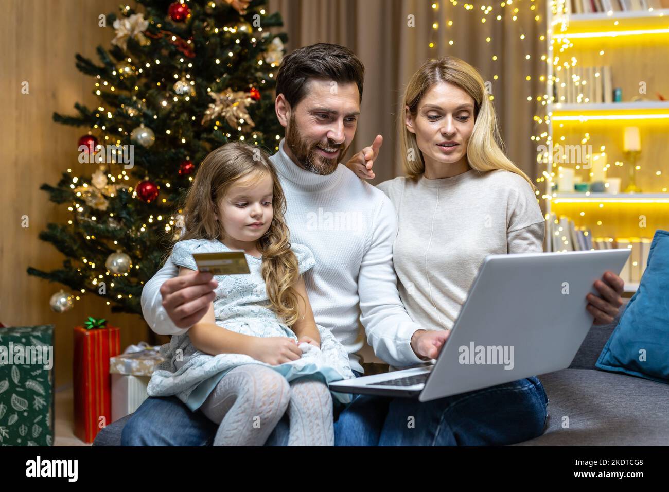 Festive online shopping. A young family, a man, a woman and a child are sitting at home near the Christmas tree. They are holding a laptop and a credit card, choosing Christmas gifts online. Stock Photo