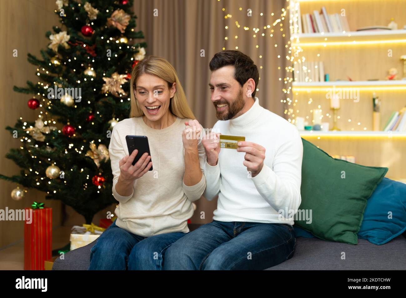 Holiday sale, online shopping. A young family, a couple, a man and a woman at home near the Christmas tree hold a phone and a credit card, rejoice at a successful profitable purchase, a discount. Stock Photo