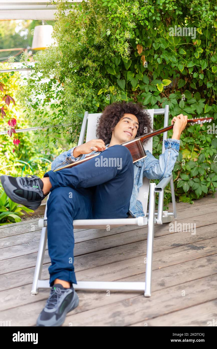 Beautiful young Afro-Italian mixed race man in casual wear playing guitar in the garden. A multiracial boy plays a musical instrument rests and Stock Photo