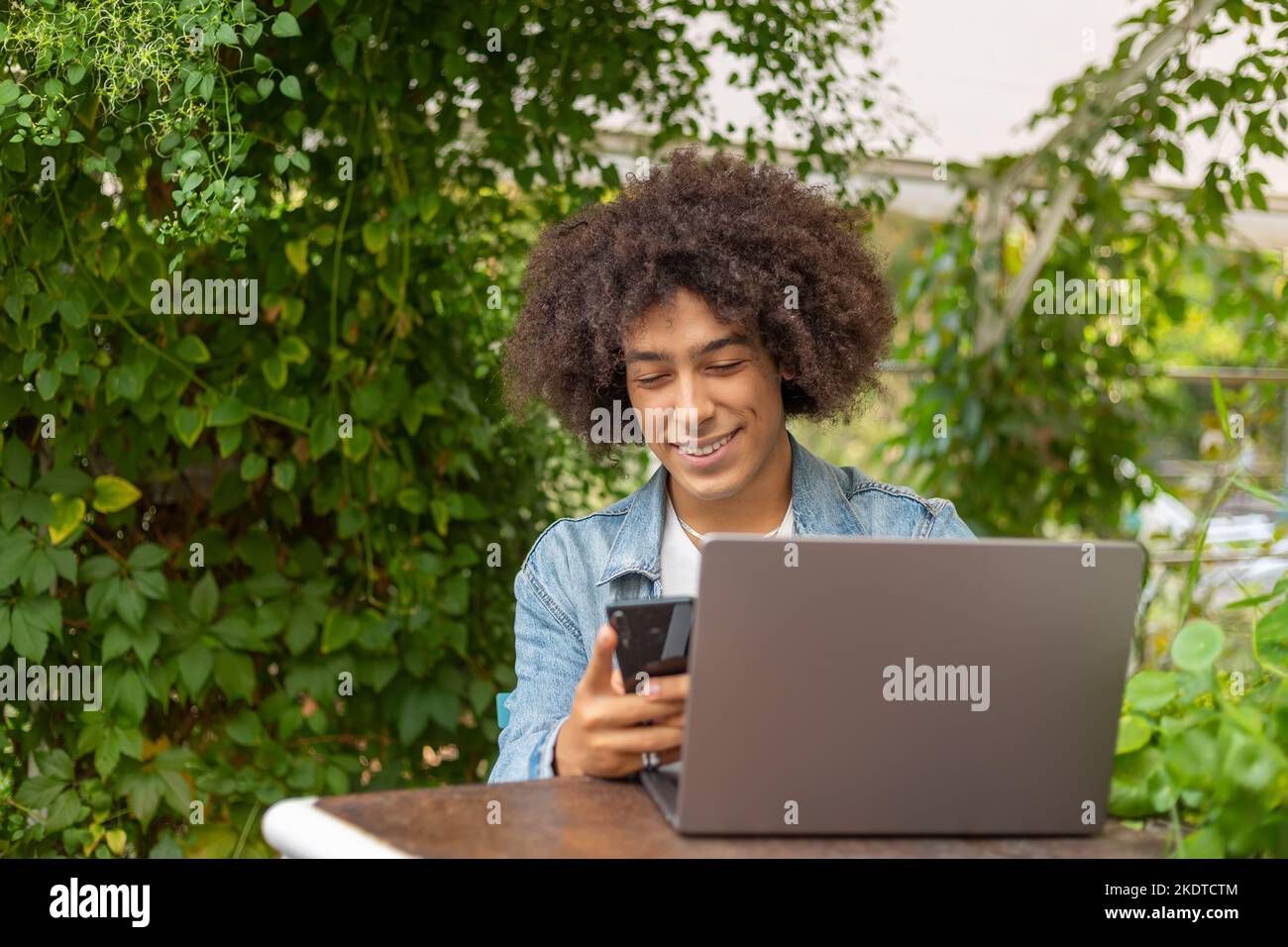 Smiling young ethnic Afro-Italian man 20 years old, dressed in casual clothes, works on a laptop outdoors, in a green area in nature. Green open space Stock Photo