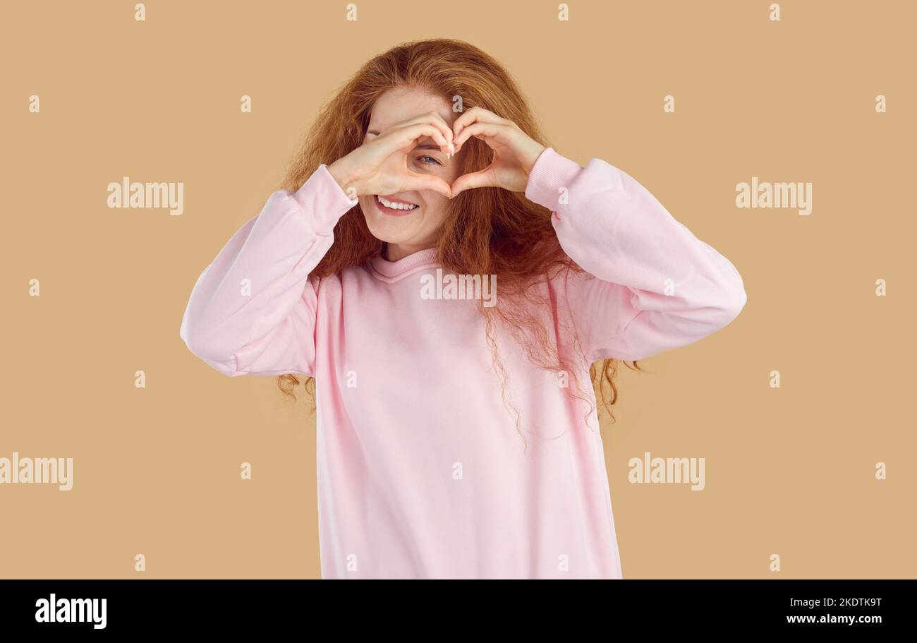 Young curly redhead girl is showing heart from hands and looking through it on beige background. Stock Photo