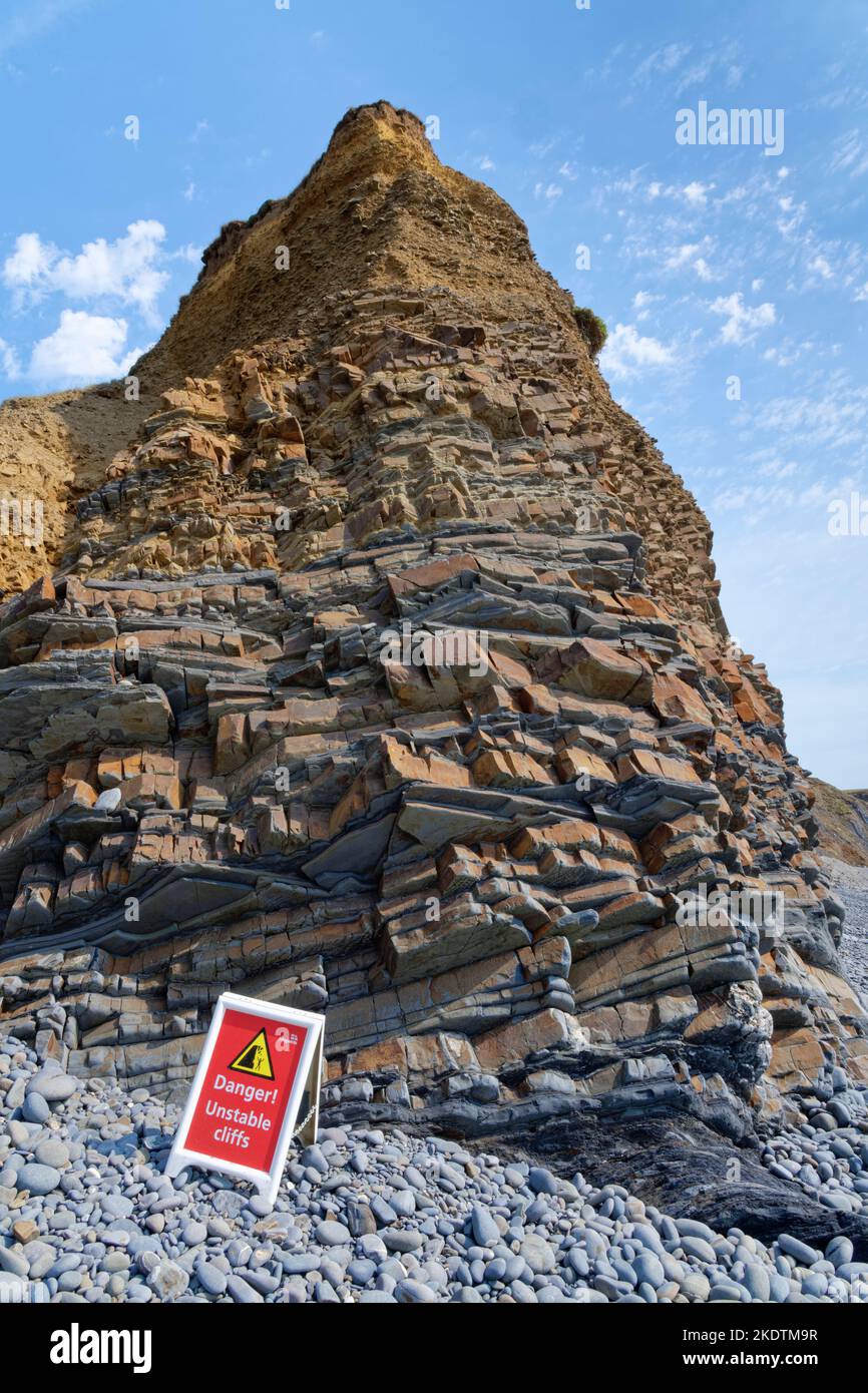 Danger of rockfall sign below unstable coastal cliffs with eroded sandstone, mudstone and siltstone rock strata, Sandymouth Bay, near Bude, north Corn Stock Photo