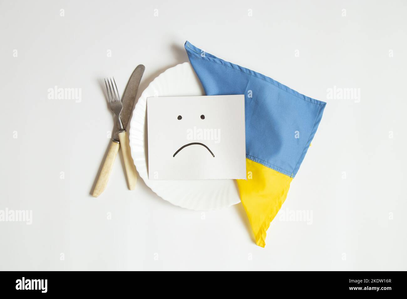 A sad emoticon is drawn on paper, lies on a plate next to the flag of Ukraine on a white background, sadness and lack of food, Ukraine without food an Stock Photo