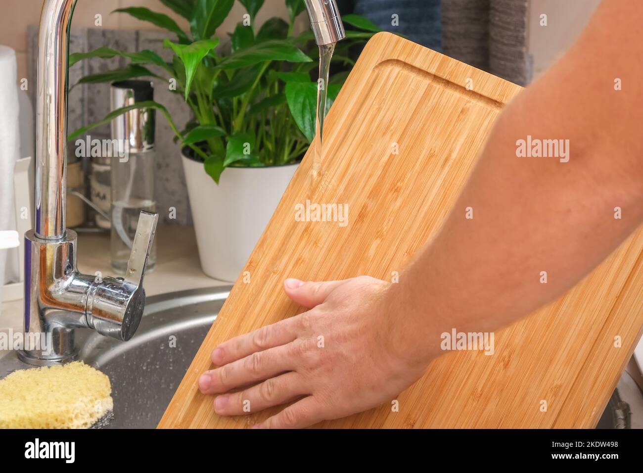 A man washes a wooden bamboo cutting board in the kitchen sink under
