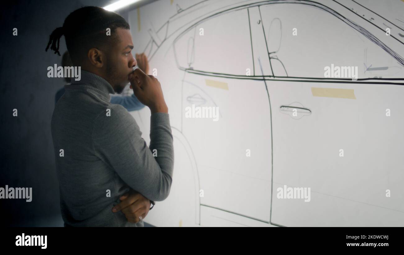 African American automotive male designer creating sketch with duct tap on whiteboard, developing new generation electric car design with coworker. Work in modern car design development studio. Stock Photo