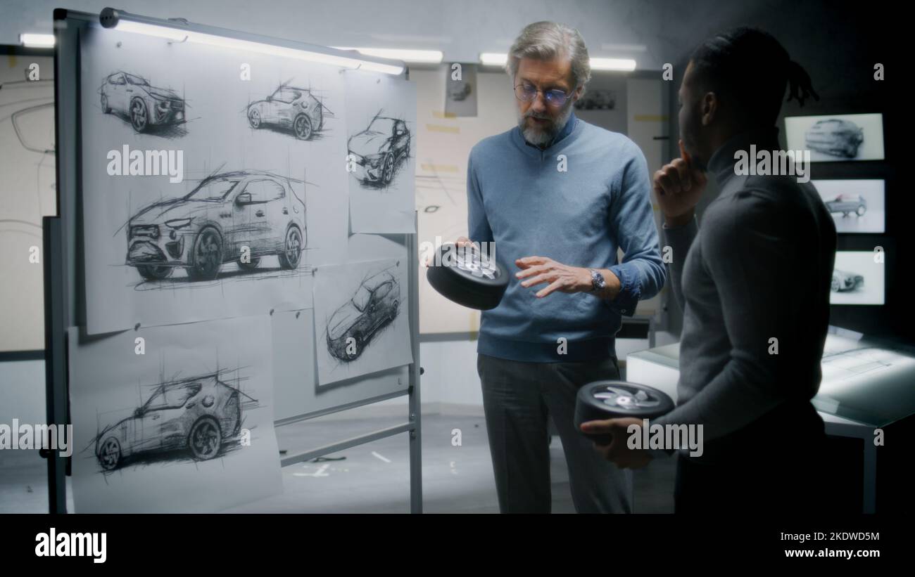 Two diverse automotive male designers holding small wheels, developing futuristic electric car design, standing near whiteboard with sketches. They working in car design development modern studio. Stock Photo